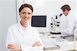 Scientist smiling at camera arms crossed and another working with microscope in laboratory