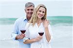 Happy couple smiling at camera and holding a glass of red wine at the beach
