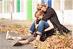 Portrait of mother and daughter sitting on curb on sidewalk in autumn, painted doors of abandoned stables in background, Vendome, Loir-et-Cher, France