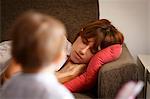 Mid adult woman sleeping  on sitting room sofa watched by toddler daughter