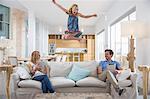 Girl jumping mid air from living room sofa whilst parents use digital tablet