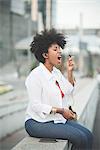 Young woman singing into earphone whilst sitting on shopping center wall