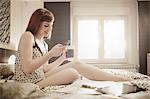 Young woman uses smart phone in bed while drinking a cup of coffee