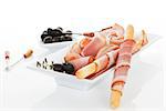 Luxurious modern minimal prosciutto background. Grissino breadsticks wrapped in prosciutto slices with black olives and fresh herbs in white tray isolated on white background. Culinary meat eating.