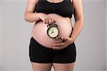 Pregnant woman showing her belly with a clock, over a gray background