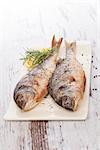 Two grilled sea bream fish on ceramic vintage cutting board on white wooden background with lemon and rosemary. Delicious healthy mediterranean seafood eating.