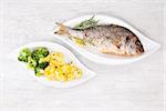 Grilled gilthead bream with fresh rosemary herb, lemon and steamed potatoes and broccoli on white plate on white wooden background. Culinary bright seafood eating.