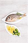 Grilled fish with steamed potatoes and broccoli, fresh herbs and lemon slice on white plate on white wooden background, top view. Culinary fish eating.