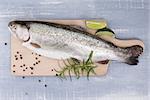 Trout on wooden kitchen board with colorful peppercorn, fresh herbs and lime on blue wooden background. Culinary seafood eating.