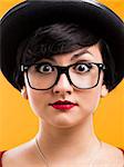 Beautiful girl with a astonished expression, wearing a hat and nerd glasses over a yellow background