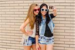 Two beautiful and young girlfriends taking pictures, in front of a brick wall