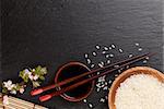 Japanese sushi chopsticks over soy sauce bowl, rice and sakura blossom on black stone background. Top view with copy space