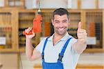 Smiling repairman with drill machine gesturing thumbs up against workshop