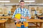 Smiling handyman holding hardhat and hammer against workshop