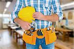 Manual worker wearing tool belt while holding hammer and helmet against workshop