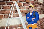 Thoughtful electrician with arms crossed by ladder against red brick wall