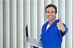 Mechanic holding laptop while showing thumbs up  against grey shutters