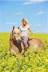 Portrait of young woman sitting on a Haflinger horse in a canola field in spring, Bavaria, Germany