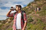 Young man at bottom of hill, drinking from flask, friends trailing behind