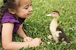 A young girl on the grass looking closely at a young duckling.