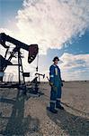 A man in overalls and hard hat at a pump jack in open ground at an oil extraction site.