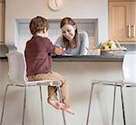 Boy sitting on stool with mother in kitchen and drawing