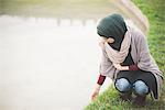 Young woman wearing hijab crouching next to park lake