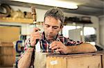 An antique furniture restorer holding a chisel and working on a polished wood surface, blowing away dust.