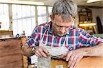 A man in a furniture restoration workshop using a hand tool on a piece of antique furniture.