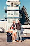 Young couple with luggage by propeller airplane, man using phone