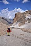 Woman dressed in red windcoat and yellow trousers climbs a mount. Mount slope is covered with snow. Blue sky with clouds and red rocks are in the background.