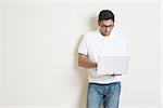 Portrait of handsome Indian guy using notebook computer, standing on plain background with shadow, copy space at side.