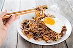 Close-up human hand holding chopsticks eating stir fried char kuey teow with over wooden background.
