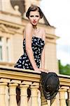 outside fashion shoot of cute brunette girl with summer elegant style posing on old balcony with short dress and stylish hat in the hand. Looking in camera.