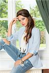 fashion portrait in morning light of cute brunette casual woman sitting near window on green garden and wearing cool jacket, modern jeans and stylish make-up