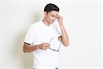 Portrait of tired Indian guy take off eyeglasses and rubbing his forehead. Asian man standing on plain background with shadow and copy space. Handsome male model.