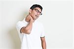 Portrait of tired Indian guy massaging his neck with painful face expression. Asian man standing on plain background with shadow and copy space. Handsome male model.
