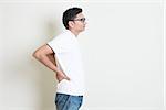 Portrait of Indian guy back pain, holding spine with hands. Asian man standing on plain background with shadow and copy space. Handsome male model.