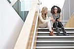 Two businesswomen sitting on office stairway reading smartphone texts