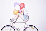 Studio shot of young woman sitting on bicycle hiding behind bunch of balloons