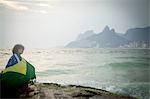 Young man wrapped in Brazilian flag sitting on rock, Ipanema beach, Rio De Janeiro, Brazil