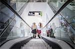 Two female friends moving up escalator in shopping mall