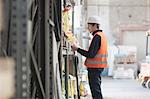 Young male warehouse worker searching shelves with clipboard in warehouse