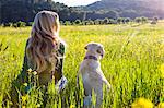 Rear view of mature woman and labrador retriever sitting in sunlit wildflower meadow