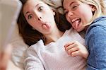 Two girls lying on bed sticking out tongues for smartphone selfie