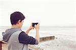 Teenage boy photographing sea on smartphone, Southend on Sea, Essex, UK