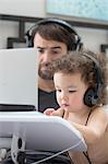 Father watching toddler daughter typing on laptop whilst wearing headphones