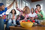 Five adult friends celebrating with high five whilst watching TV from sofa