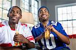 Two male friends drinking bottled beer whilst watching TV from sofa
