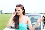 Frustrated woman using cell phone against broken down car